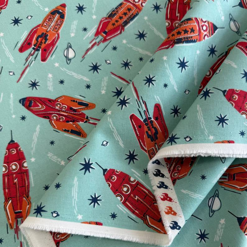 Retro red and orange rockets flying across a light aqua blue background with stars and planets, Blast Off Blue Nebula quilting cotton by Stacy Iest Hsu for Moda Fabrics.