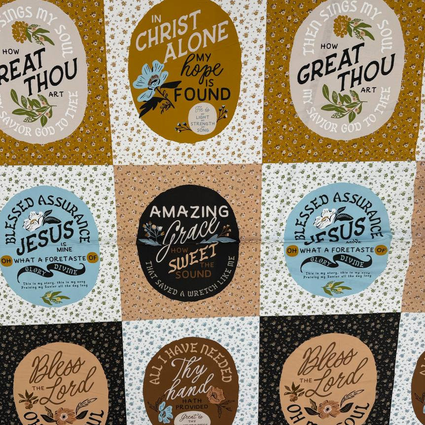 A square patchwork-style fabric or mat with a grid of circular and oval panels in black, cream, tan, light blue, and mustard yellow, each printed with decorative text and small illustrations. The product is photographed from above on a white tabletop, with a small plant and shoes partially visible around the edges.