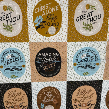 A square patchwork-style fabric or mat with a grid of circular and oval panels in black, cream, tan, light blue, and mustard yellow, each printed with decorative text and small illustrations. The product is photographed from above on a white tabletop, with a small plant and shoes partially visible around the edges.