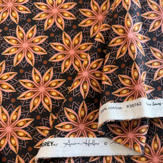 Acorn Hollow quilting cotton fabric by BasicGrey for Moda Fabrics featuring repeating geometric starburst motifs in pink, gold, and amber tones on a deep soil brown background.