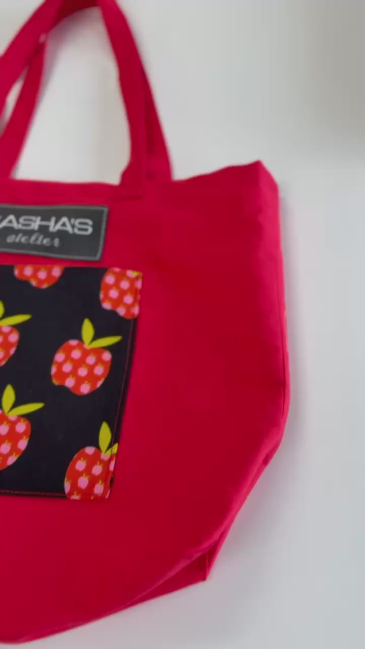 Handmade red canvas tote bag with two exterior pockets (front and back), featuring a decorative apple-print front pocket, comfortable shoulder straps, and Sasha's Atelier label. Proudly made in Canada for shopping, work, travel, and everyday use.
